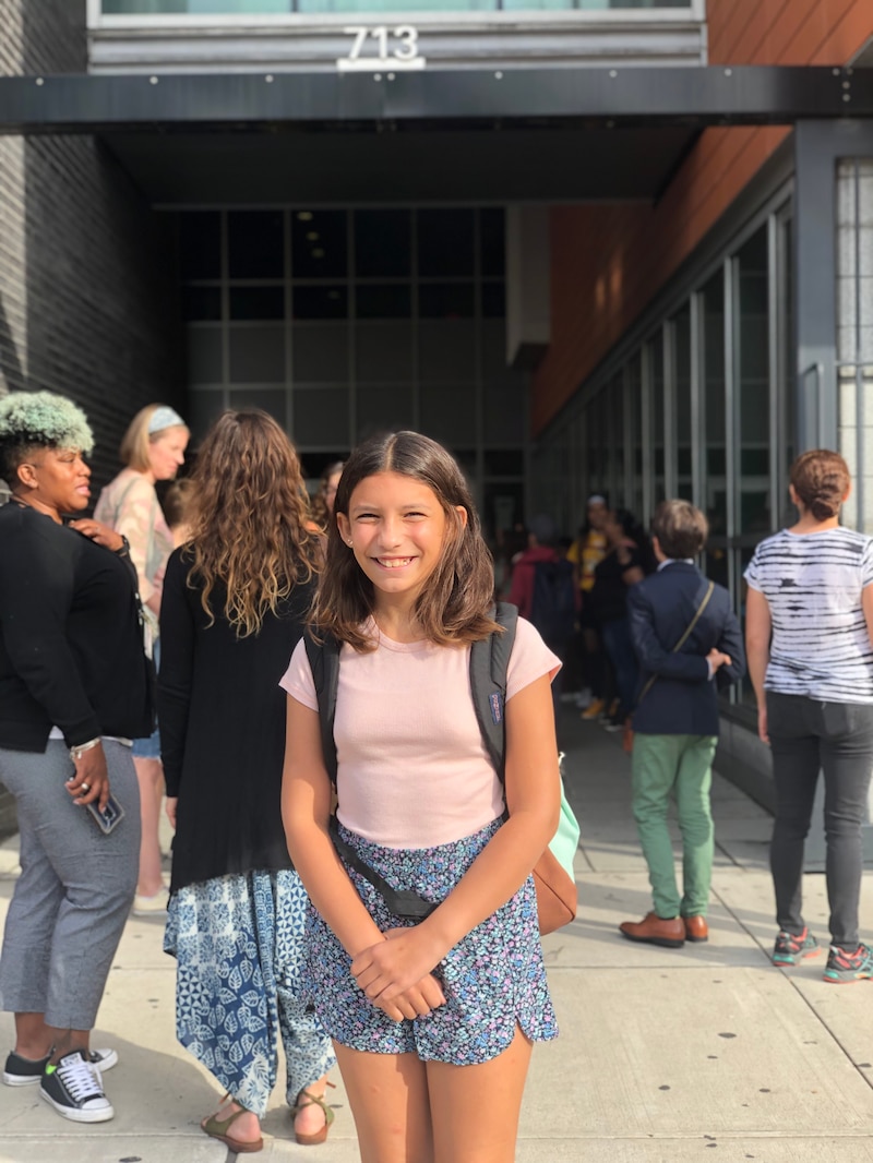 Photo of a younger girl wearing a pink tee shirt and blue shorts. She is carrying a backpack.
