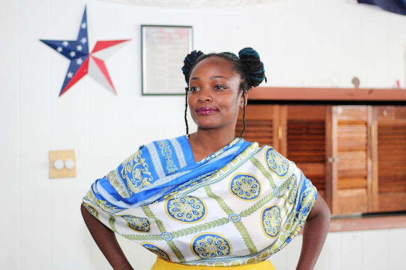 A woman wearing a patterned blouse poses for a portrait in front of a wall with an American flag star and a wooden shelf.