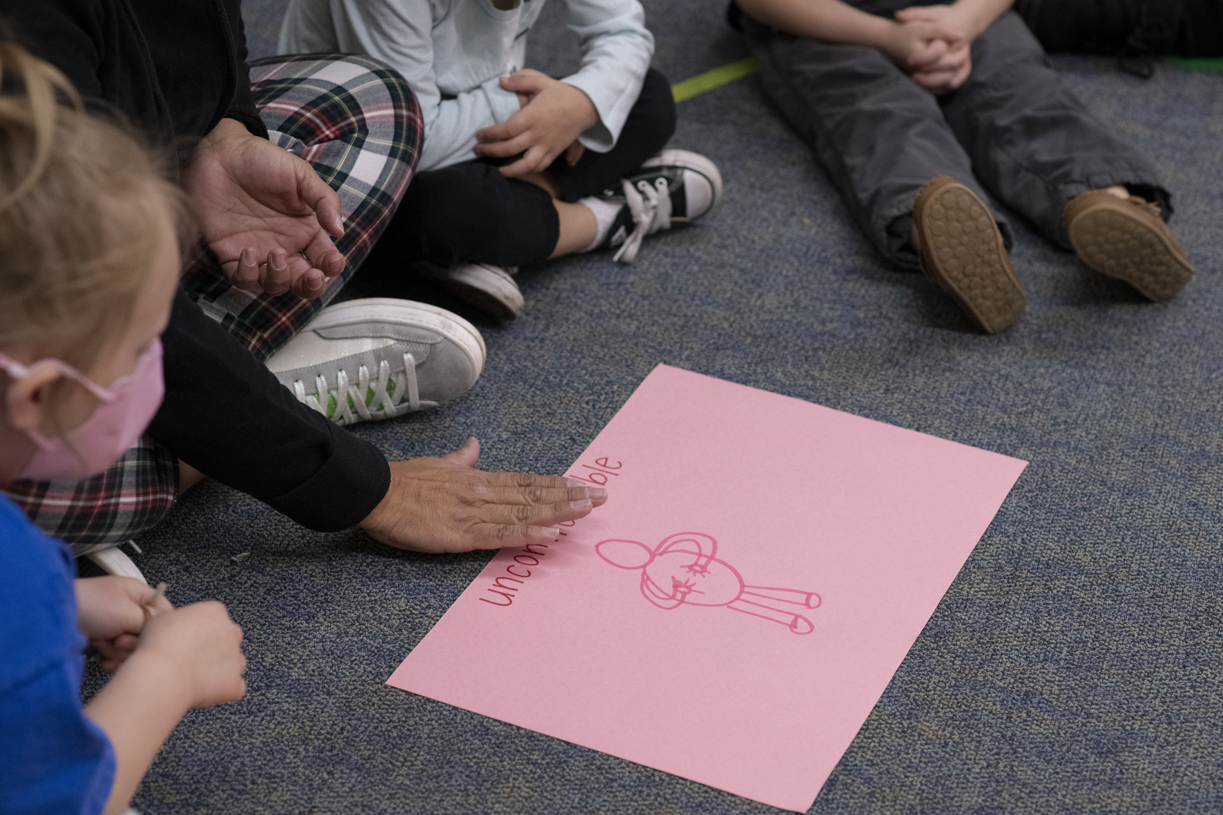 A teacher points to a piece of pink construction paper that has a stick figure drawing and the word “uncomfortable” on it. The teacher is sitting on the rug. Only her feet and hands are visible.