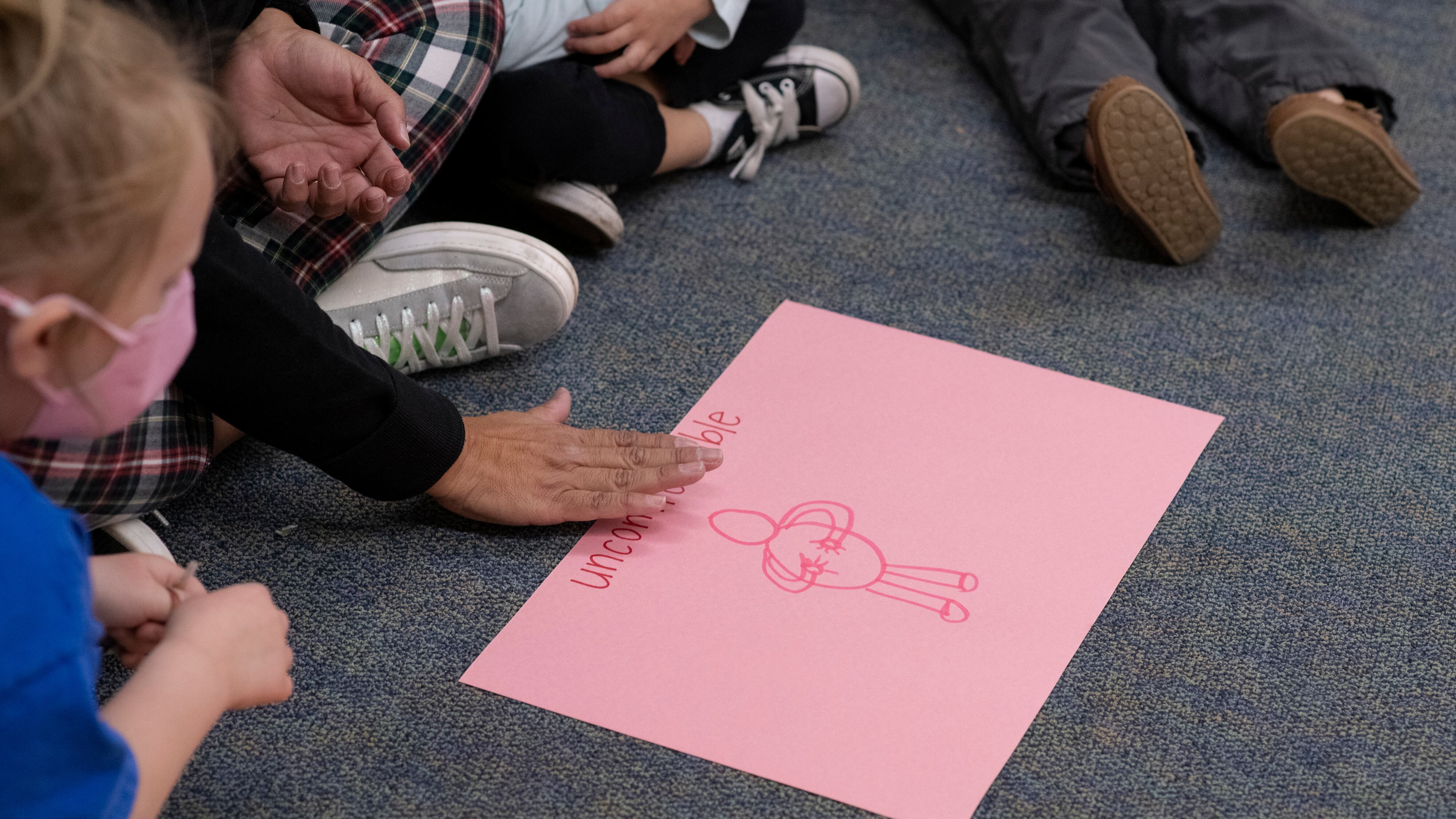A teacher points to a piece of pink construction paper that has a stick figure drawing and the word “uncomfortable” on it. The teacher is sitting on the rug. Only her feet and hands are visible.