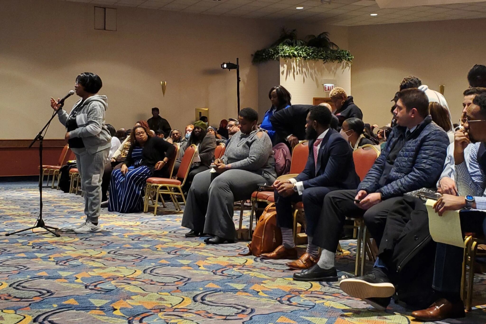 A woman wearing a gray hoodie stands in front of a microphone. Behind her are rows of men and women sitting in chairs. The carpet beneath them is swirly and colorful.