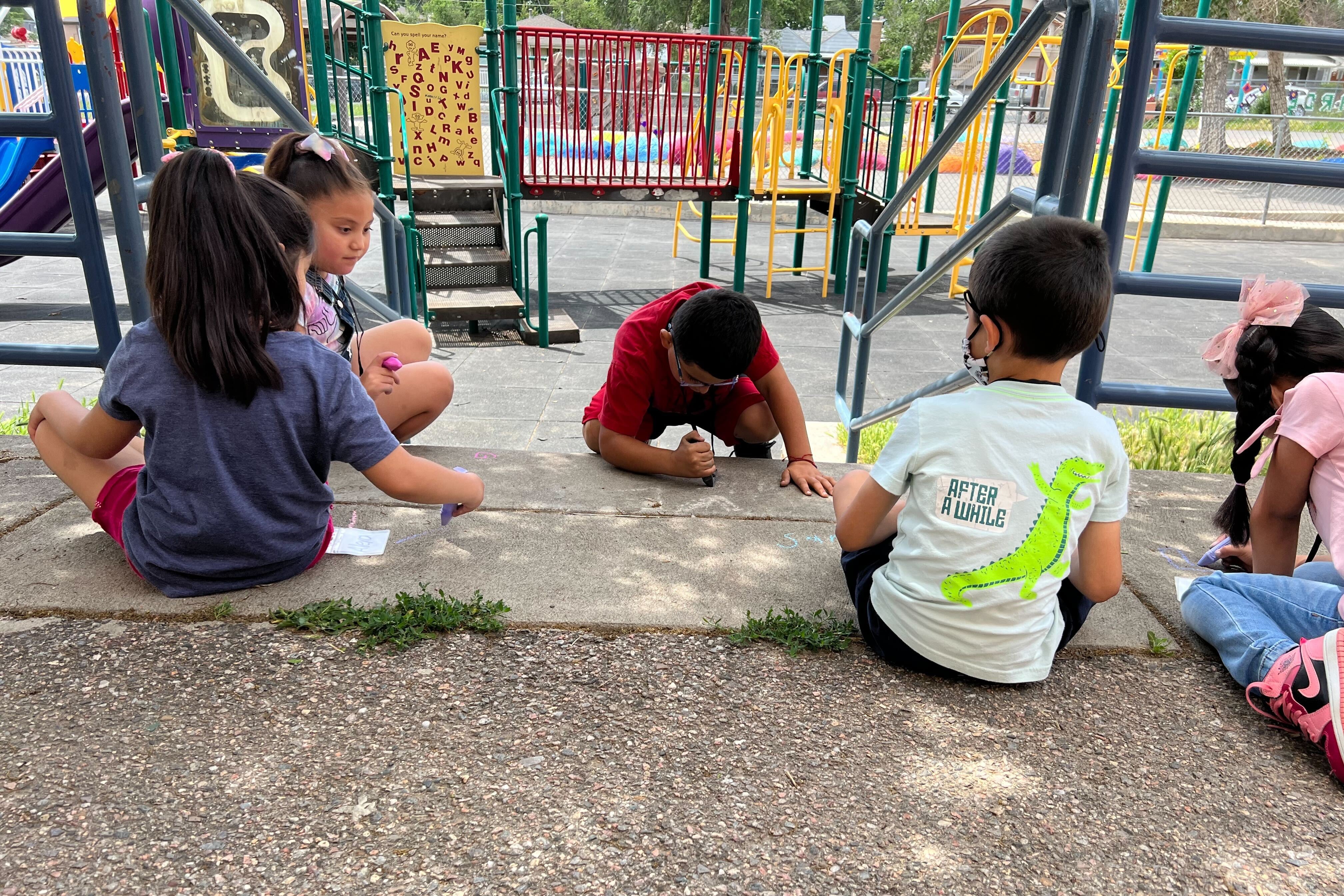 Five young children write with chalk on the sidewalk in front of a school playground.