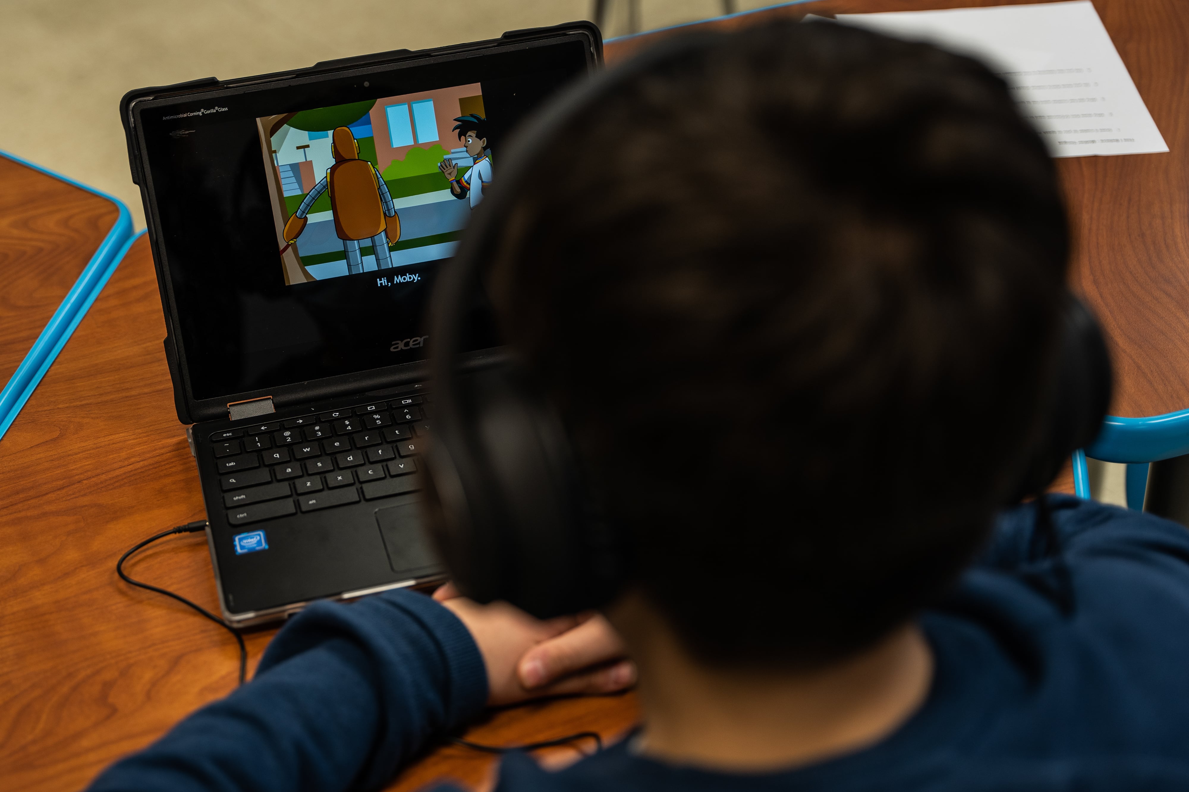A child with dark hair and a blue shirt wearing headphones watches a screen on a laptop while sitting.