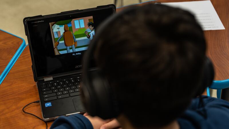 A child with dark hair and a blue shirt wearing headphones watches a screen on a laptop while sitting.