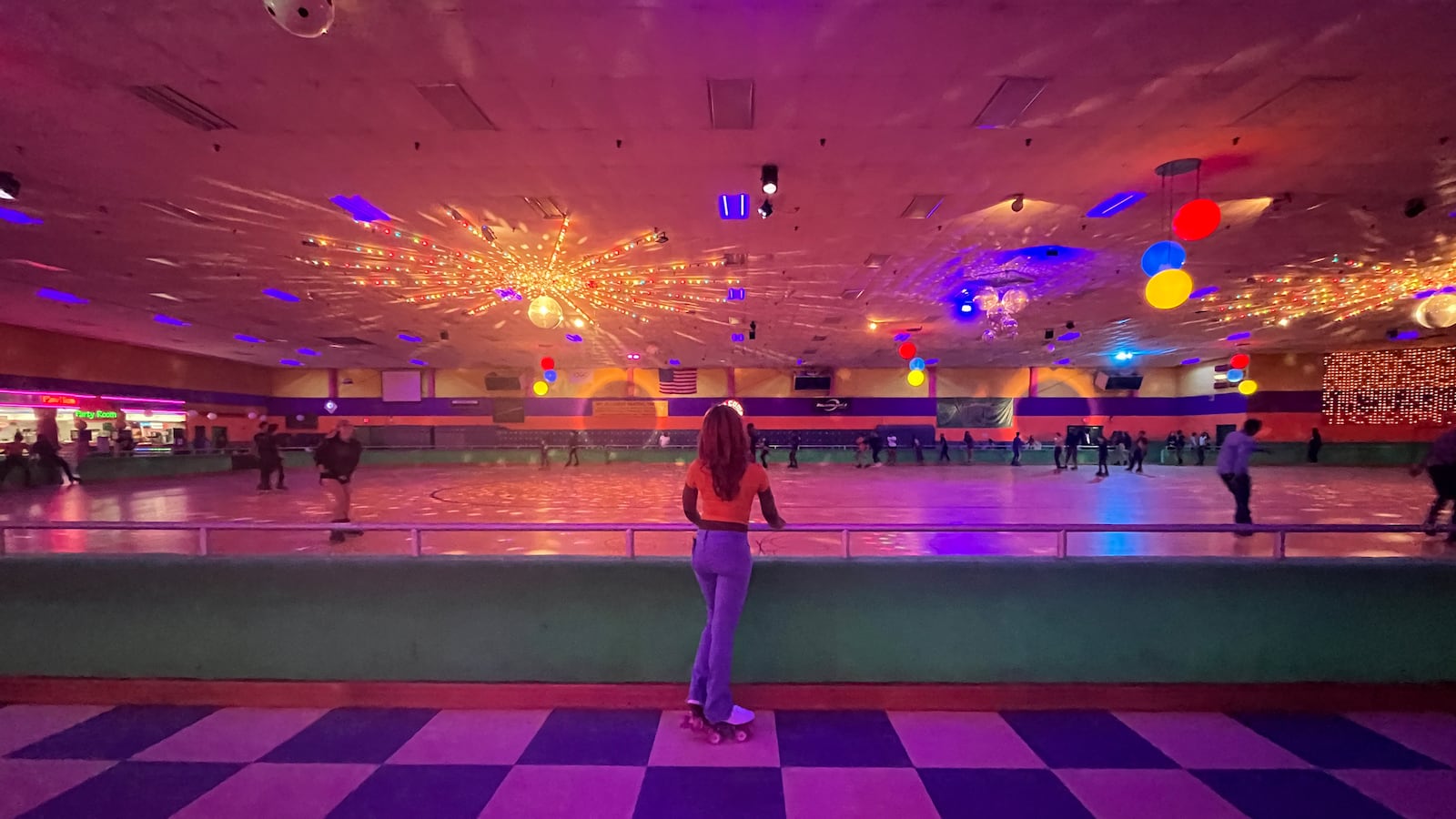 A high school girl wearing roller skates stands next to a railing on a checkered floor next to a roller rink with disco balls and lots of different color lights.