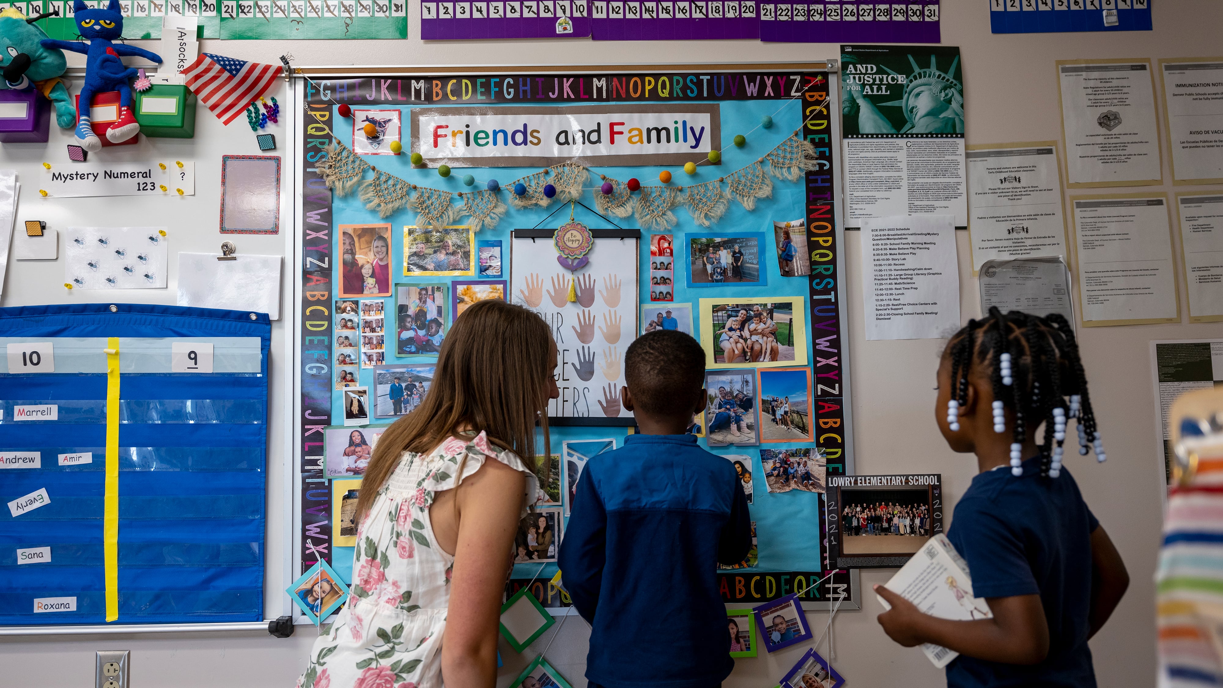 A teacher, facing a bulletin board, talks with two young students.