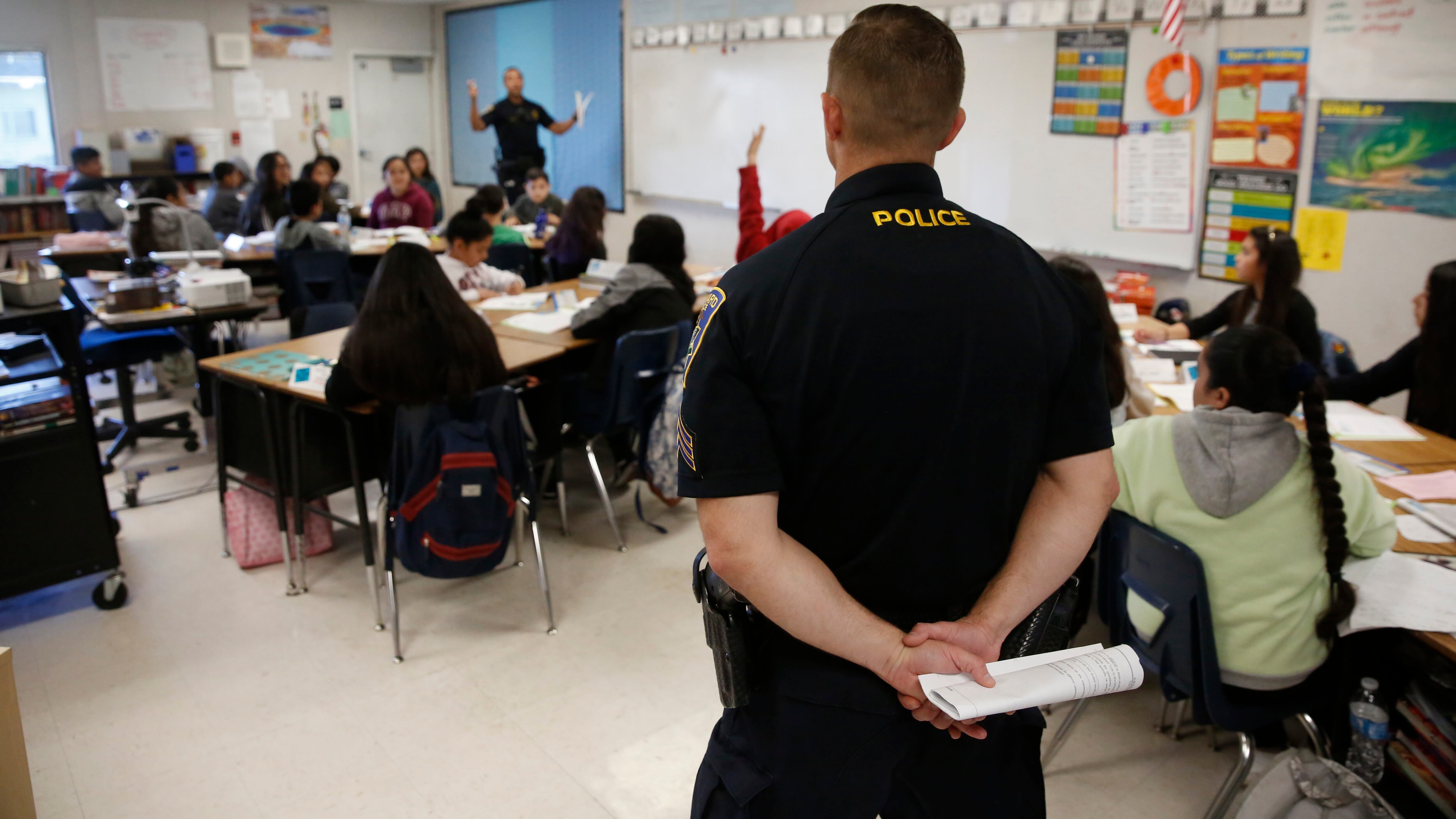 A police officer stands at the edge of a classroom, where several students sit at their desks as a teacher conducts a lesson for the students.