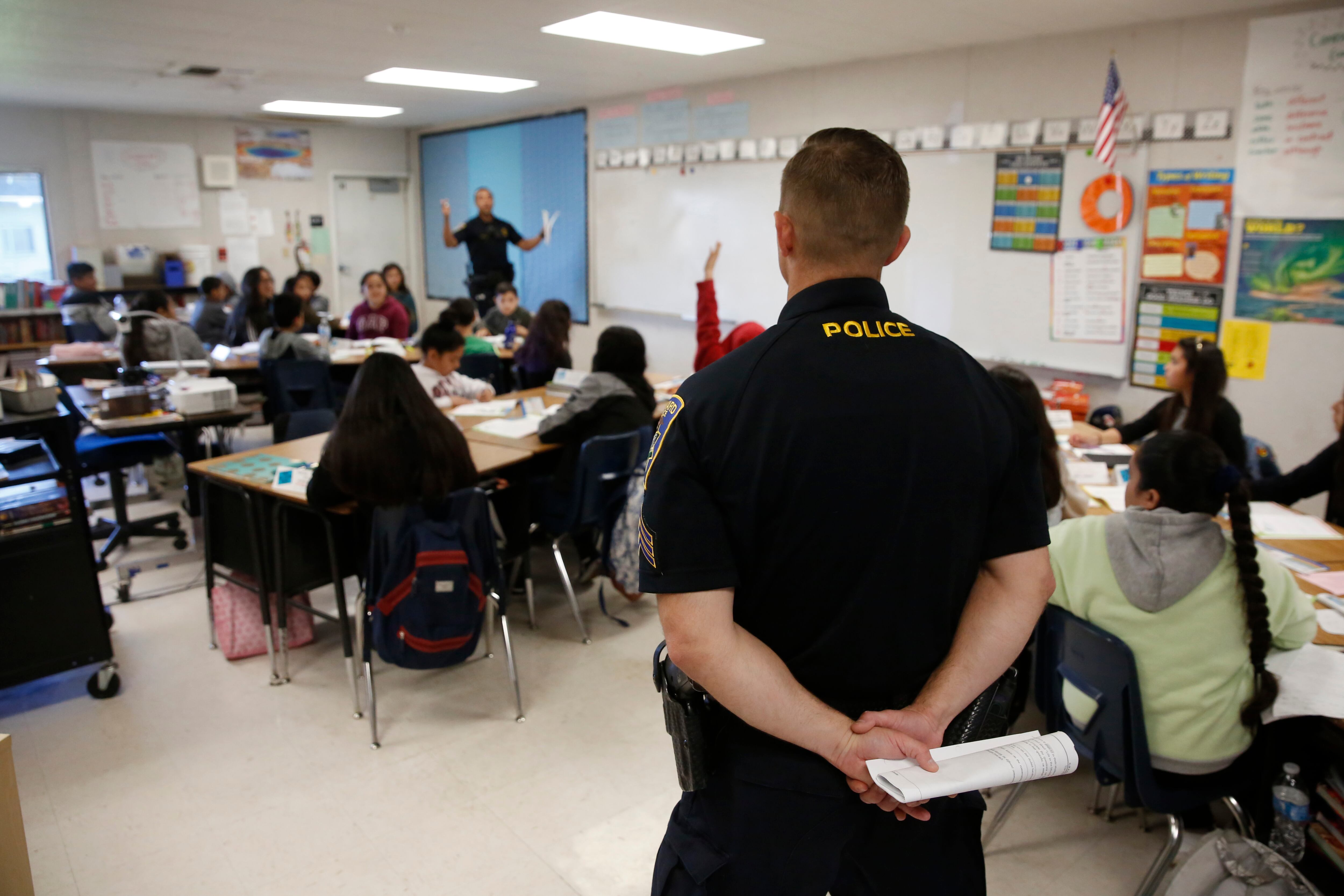 A police officer stands at the edge of a classroom, where several students sit at their desks as a teacher conducts a lesson for the students.
