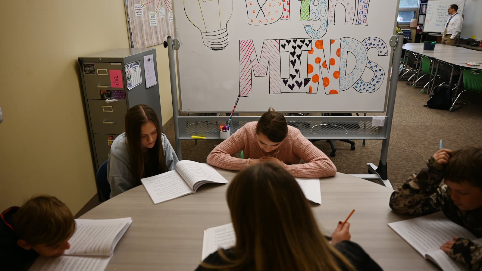 Four teenage students look down at workbooks, along with an adult holding a pencil, all sitting at a circular table. A whiteboard behind one of the students reads “Bright MINDS” in stylized, colorful letters with a lightbulb drawn to the left.