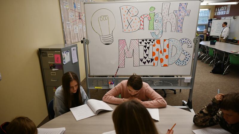 Four teenage students look down at workbooks, along with an adult holding a pencil, all sitting at a circular table. A whiteboard behind one of the students reads “Bright MINDS” in stylized, colorful letters with a lightbulb drawn to the left.