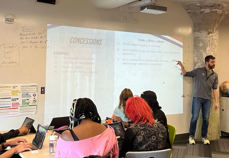 A teacher points at a projected image on a wall of a classroom with students sitting in chairs in the foreground.