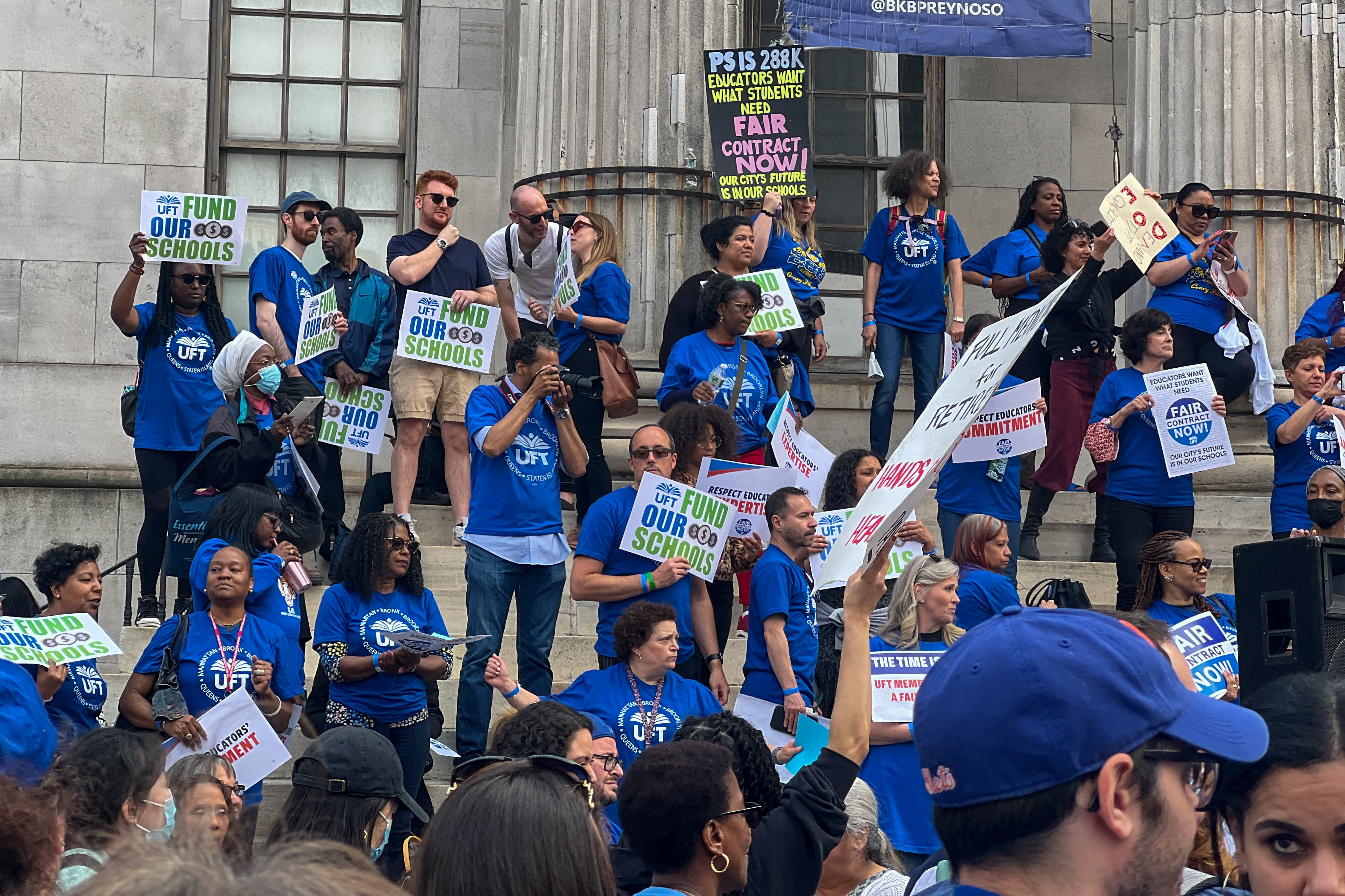 A group of people wearing bright blue T-shirts stand on the steps holding signs for a fair contract.