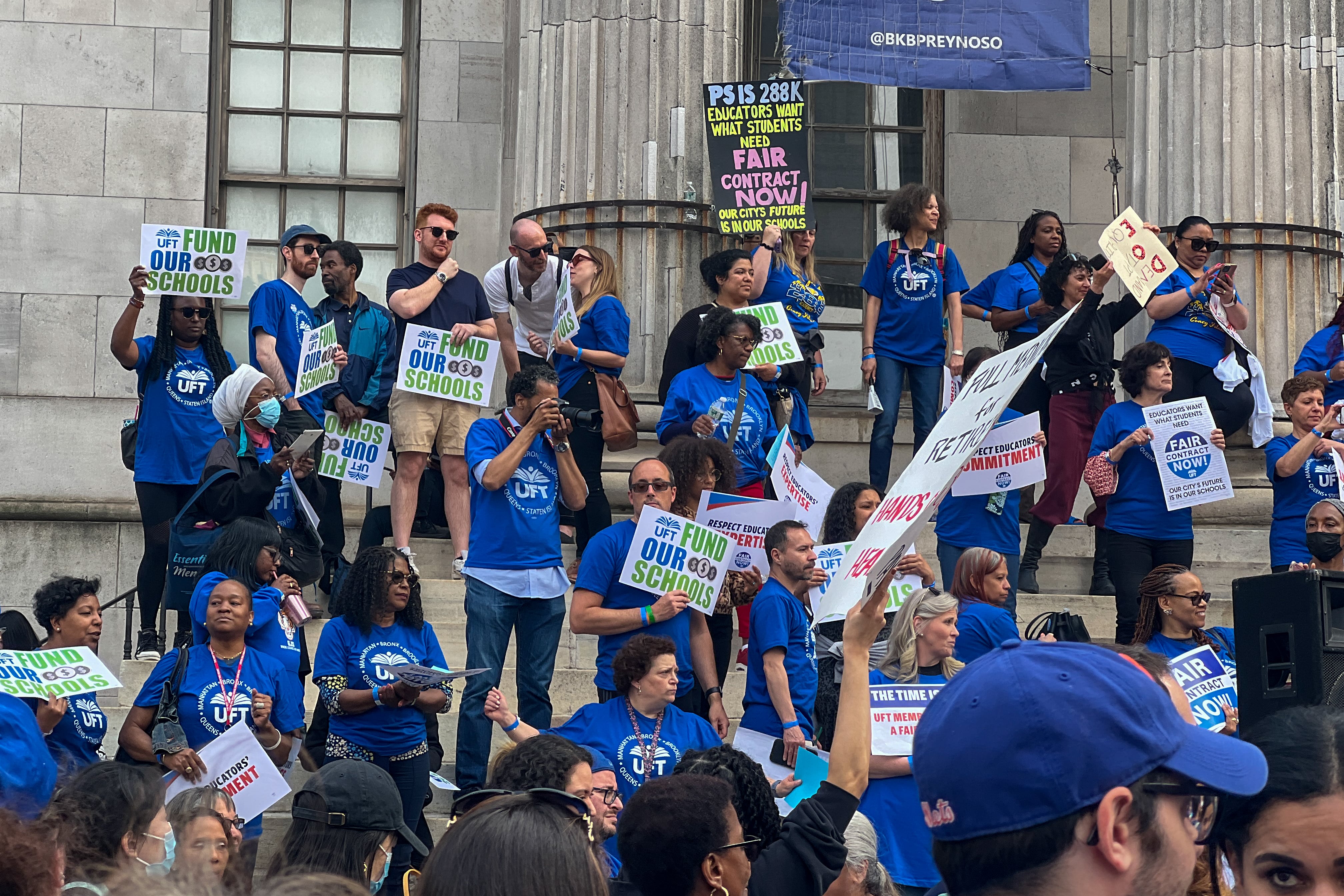 A group of people wearing bright blue T-shirts stand on the steps holding signs for a fair contract. 