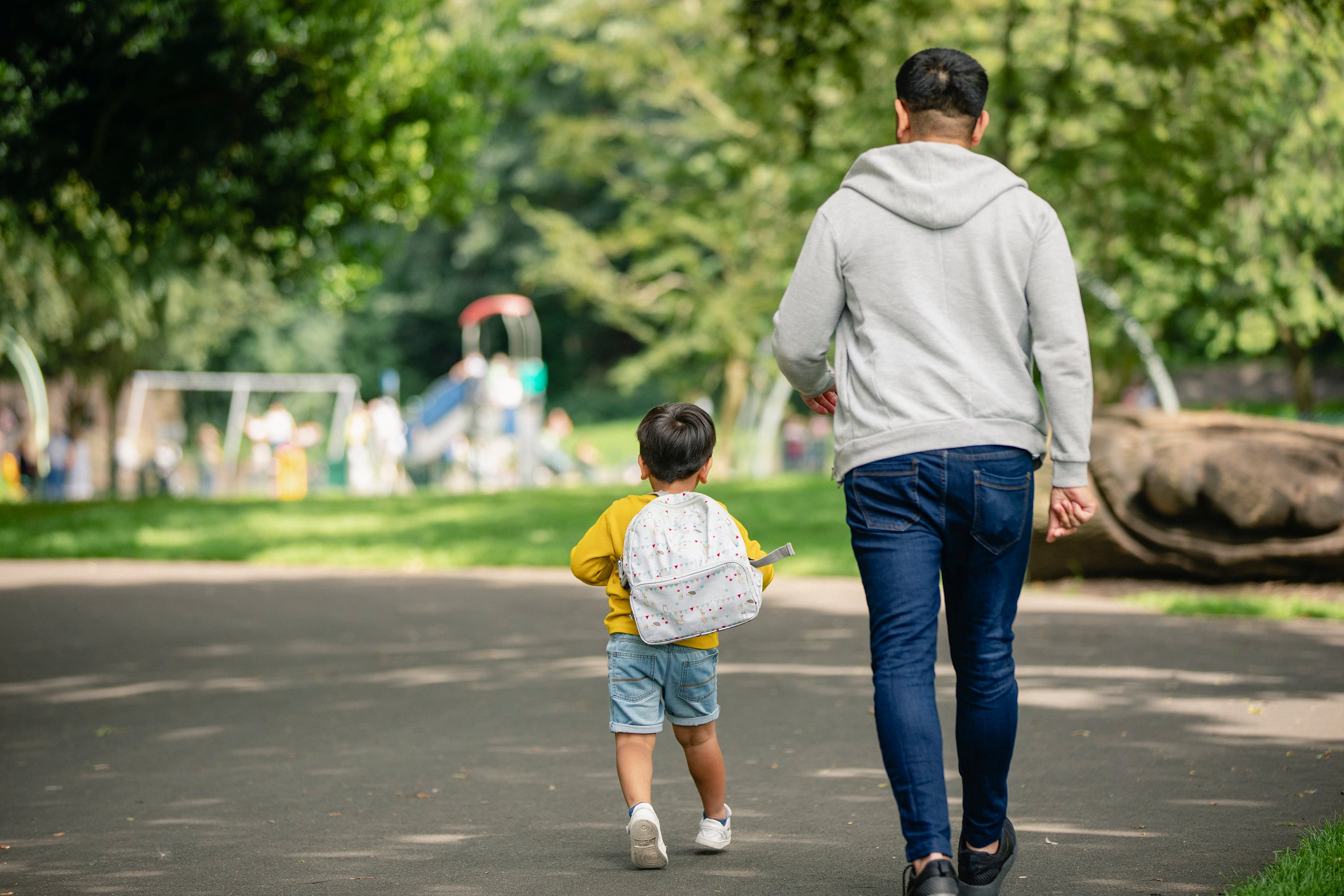 A young child wearing a small backpack walks beside an adult wearing jeans and a grey sweater outside with a green area in the background.