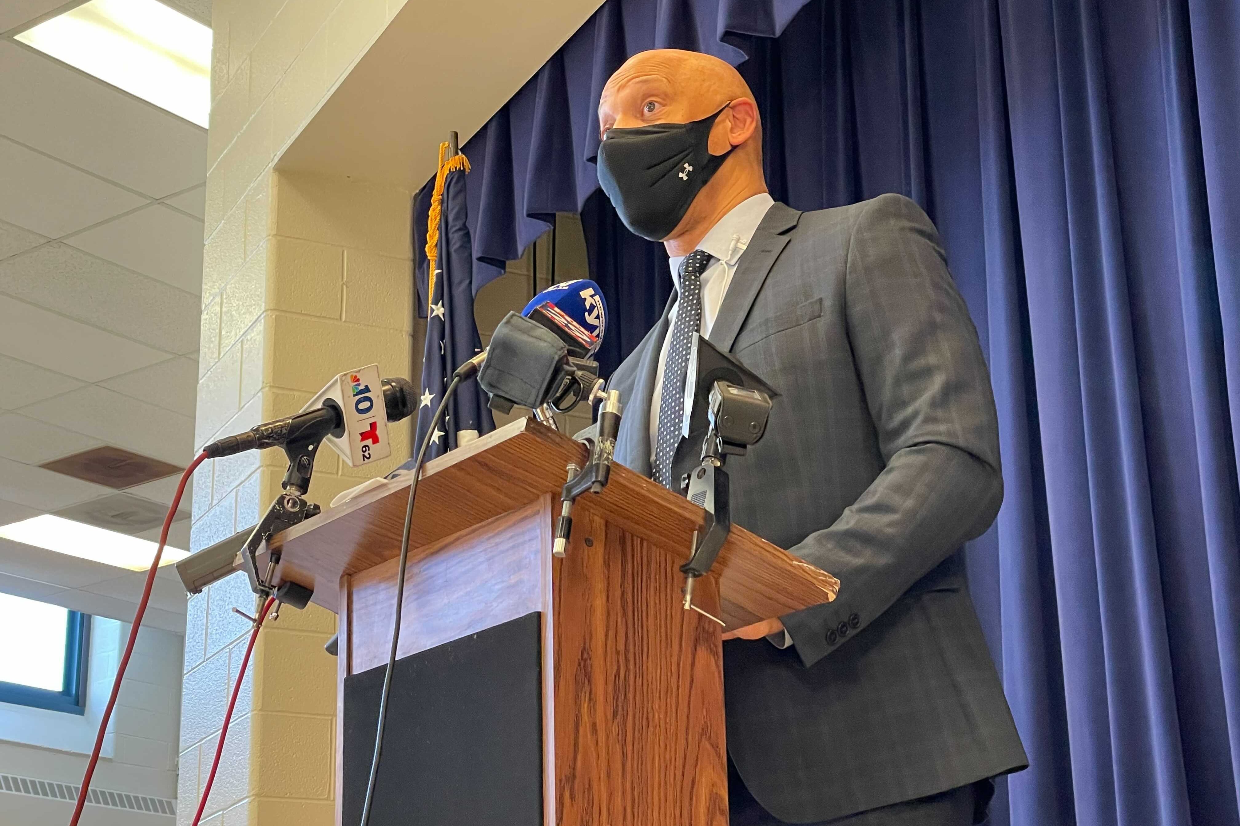 William Hite wearing a face mask standing at a podium with microphones.