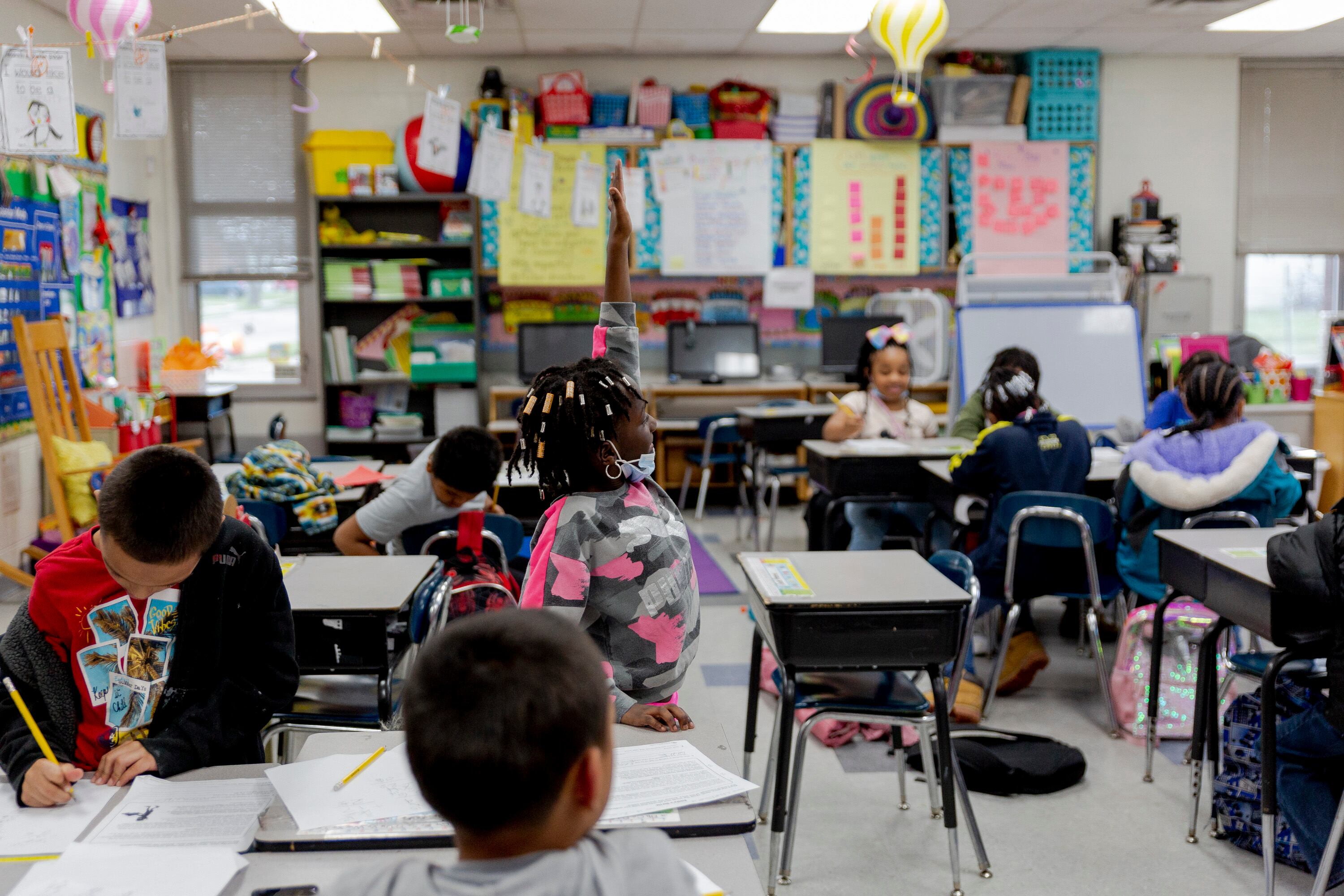A student raises her hand and stands at her desk, as several other young students work around her in the classroom.