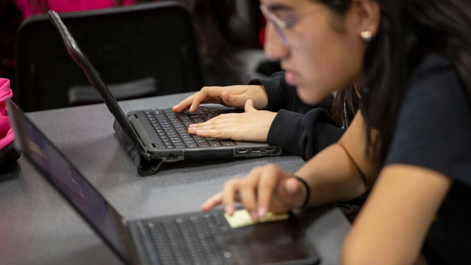 A high school student at DCIS Montbello leans toward a laptop on a table and looks intently into the screen. The hands of another student sitting to the right are seen on a laptop. The students were working on an assignment in class in May 2019.