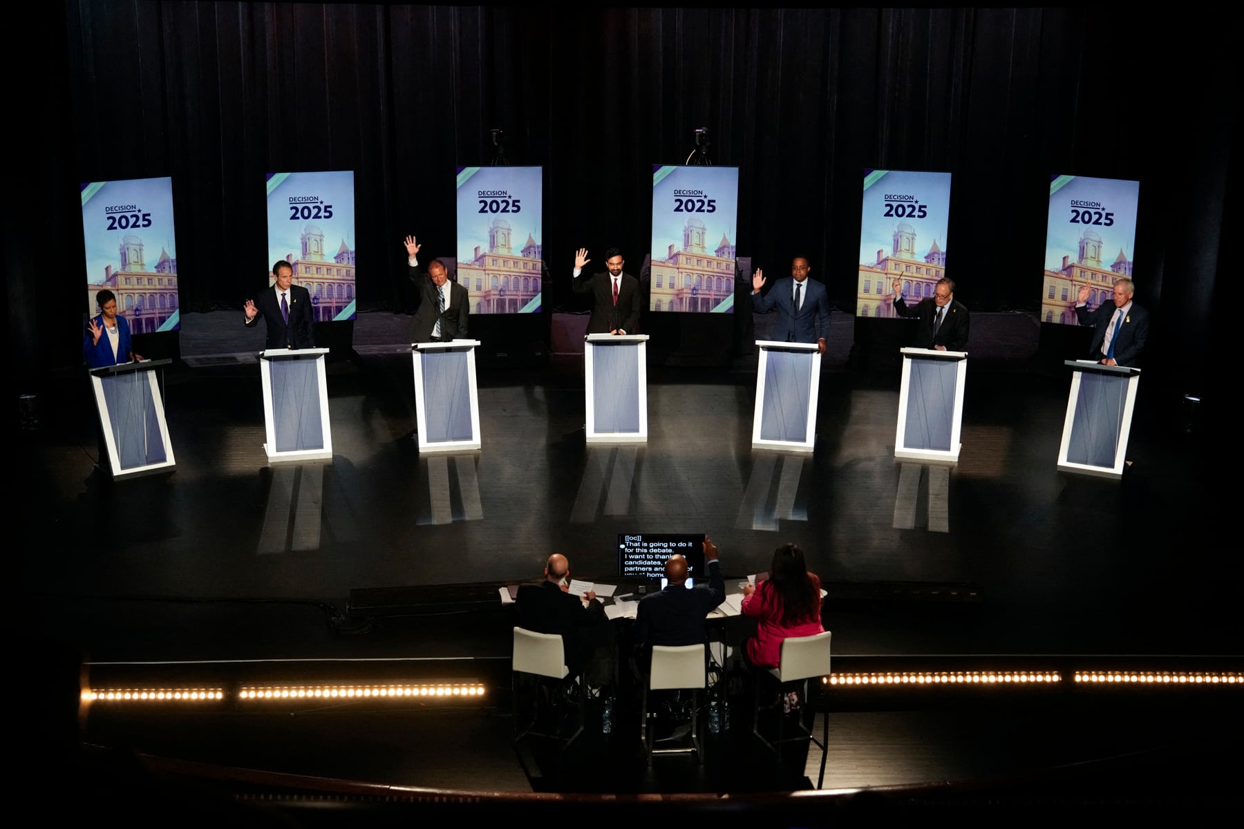 A photo of seven people in suits raise their right hand while standing behind a podium on a large stage for a debate in front of three people sitting at a small table with a screen.