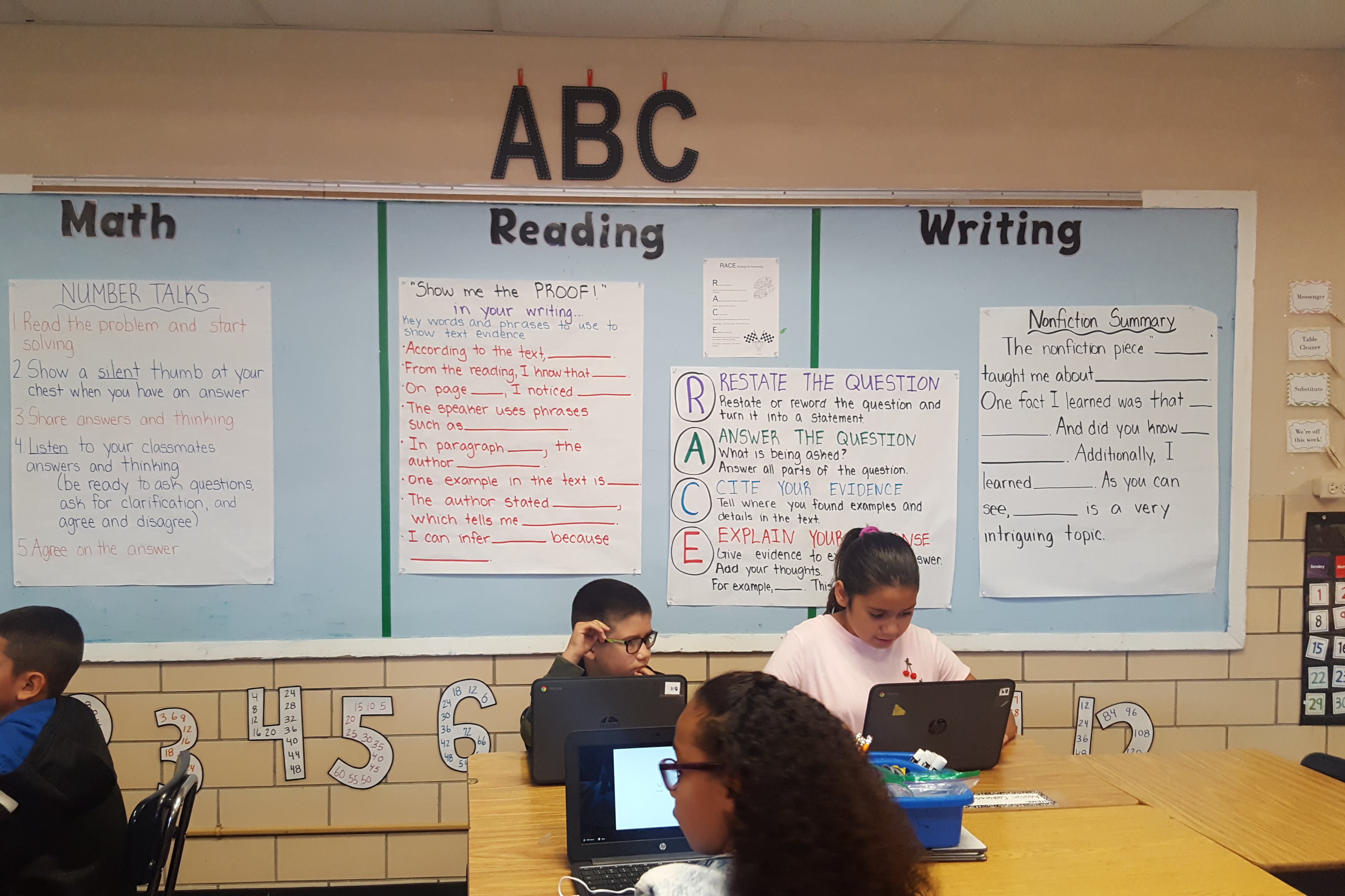 Four young elementary school students work from their desks in a classroom with math, reading and writing posters on the back wall.