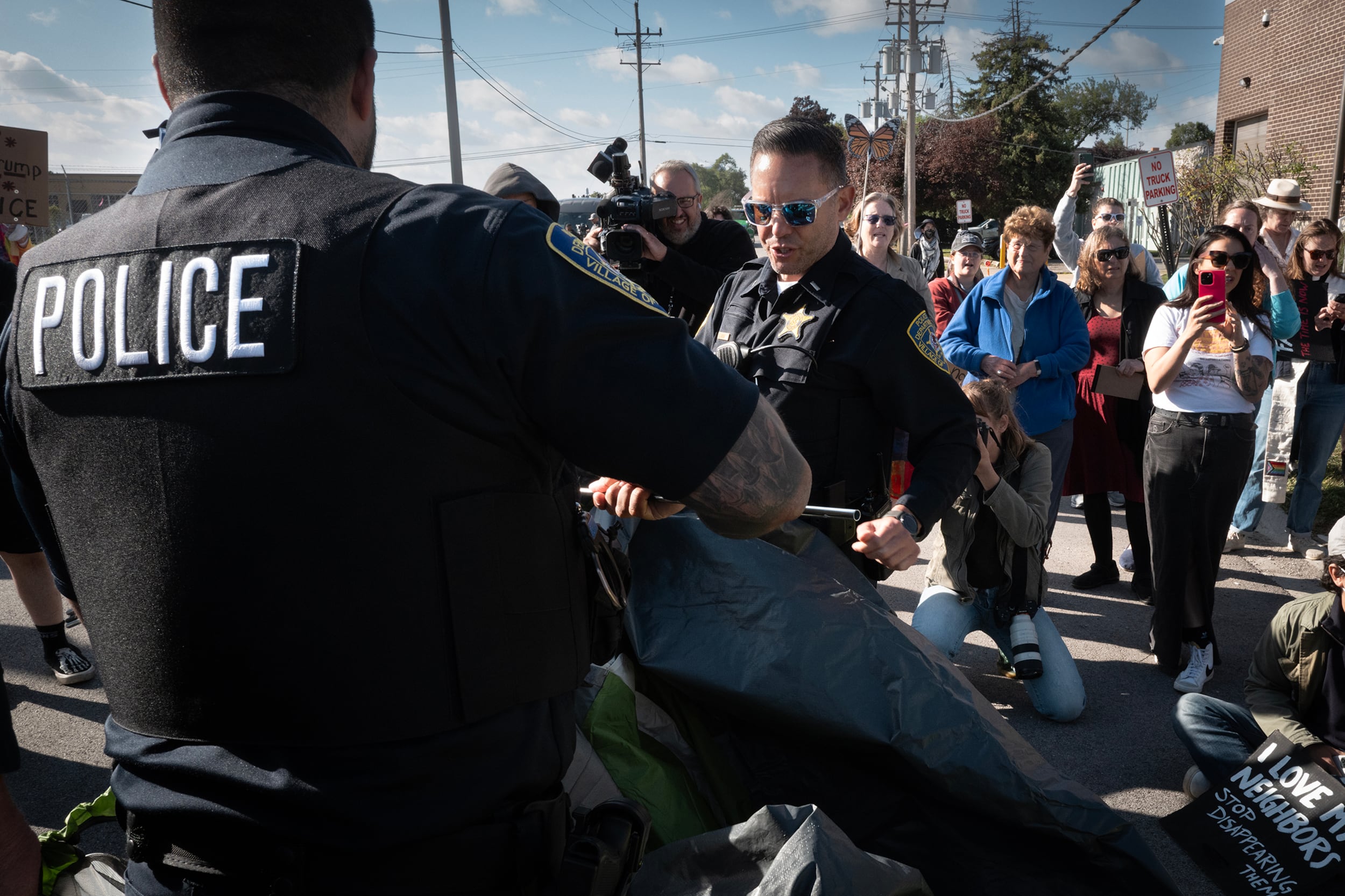 A photograph of two police officers dismantle a tent in front of a group of protestors outside on a sunny day,.