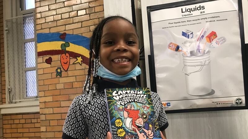 A little girl with braided hair smiles at the camera, posing with a copy of a Captain Underpants children’s book.