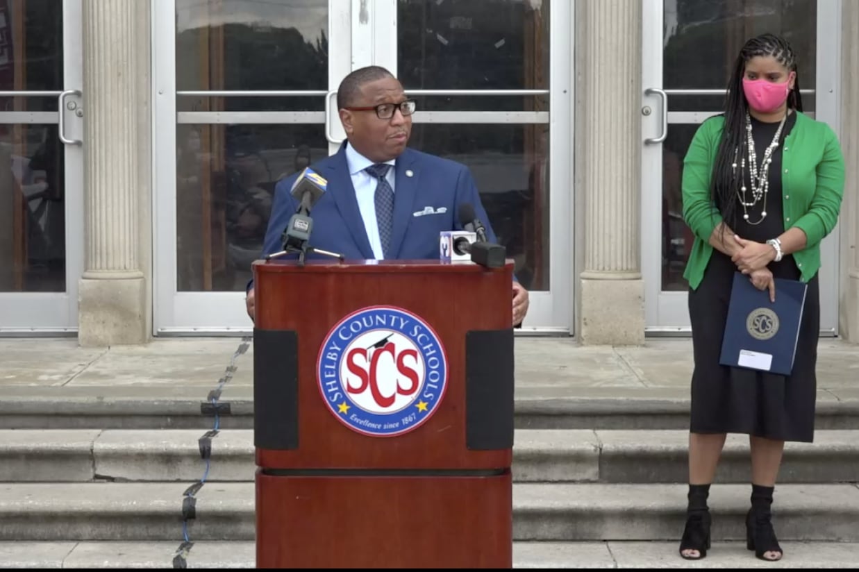 A man wearing a blue suit speaks at a podium, as a woman wearing a green blazer looks on behind him on his left side