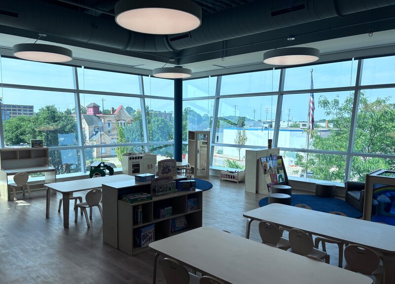 A photograph of a early childhood center room with long tables and some chairs and two bookshelves with two walls of windows in the background.