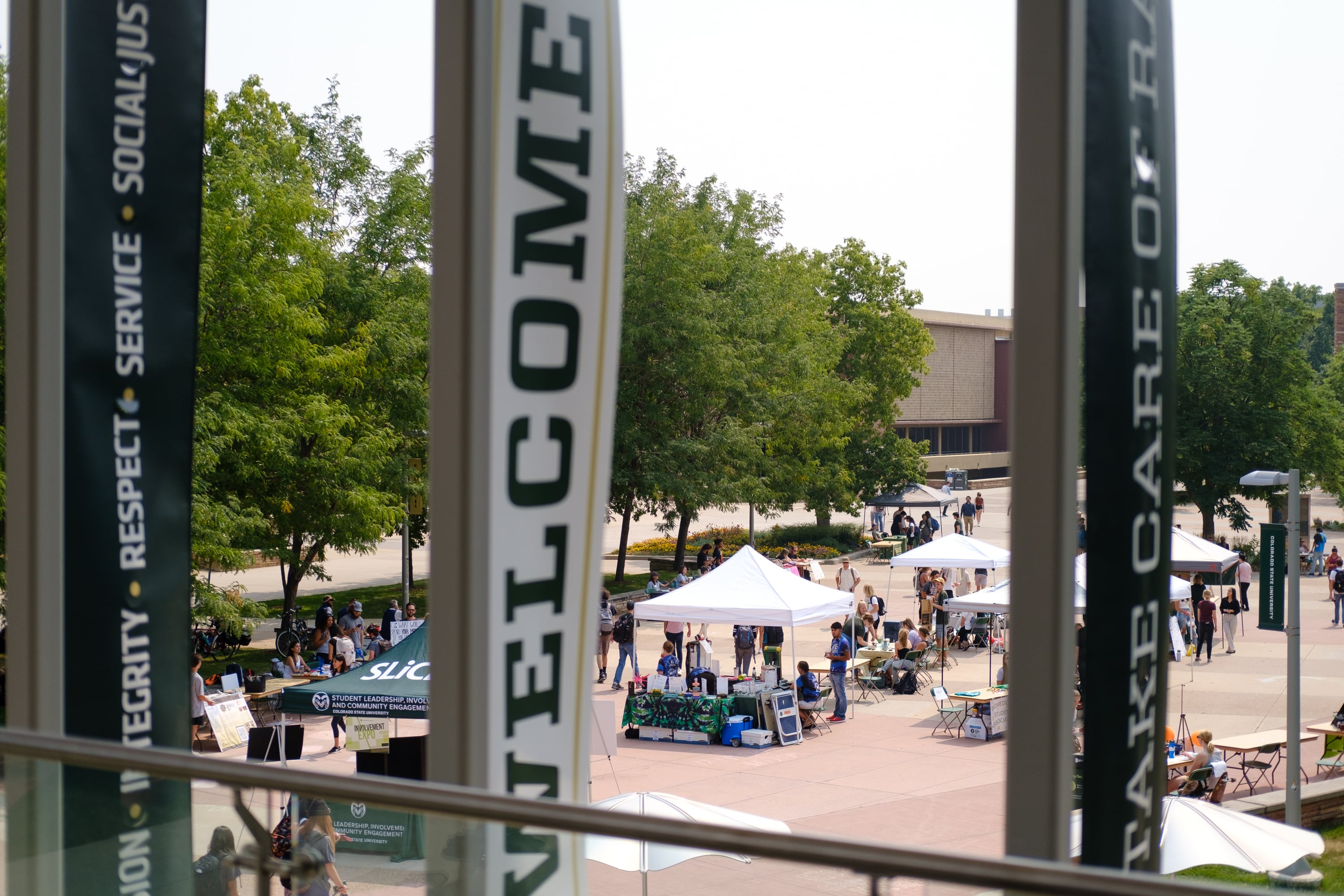 A vertical white welcome sign sits in the foreground, wotj a college campus square filled with white canopies, trees, and students in the background.