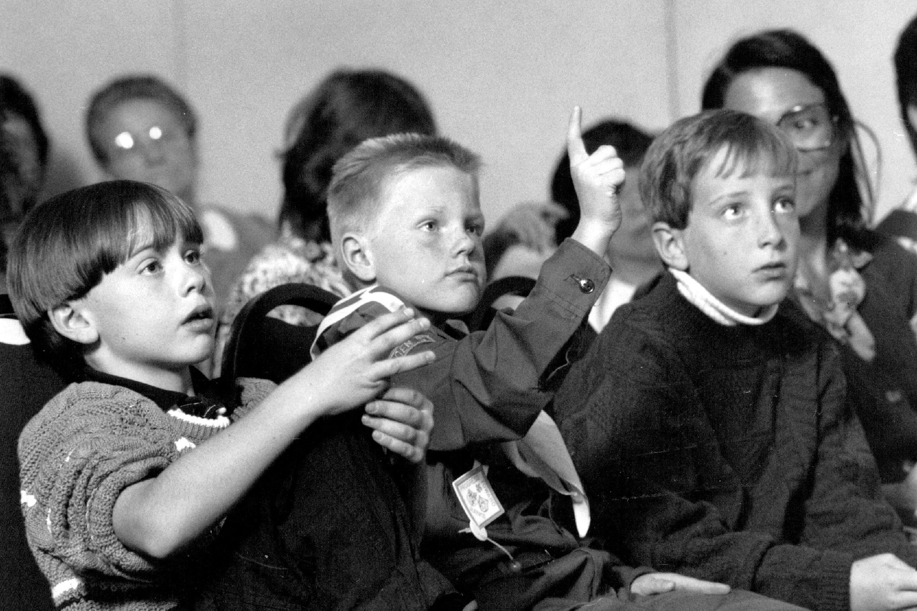 Three young students with short light hair sit in front of other students listening to someone speak.