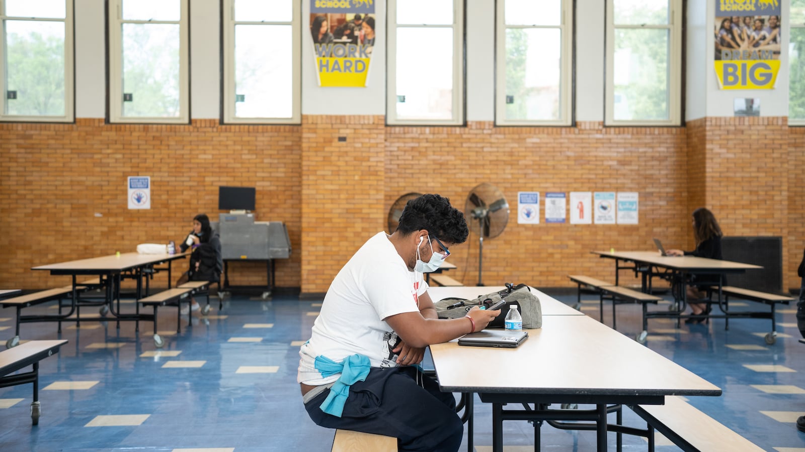 Nathaniel Martinez sits alone at a long cafeteria table. The large room, with blue tiled floors, brick walls and long windows, appears largely empty, with two students sitting by themselves at tables in the background.