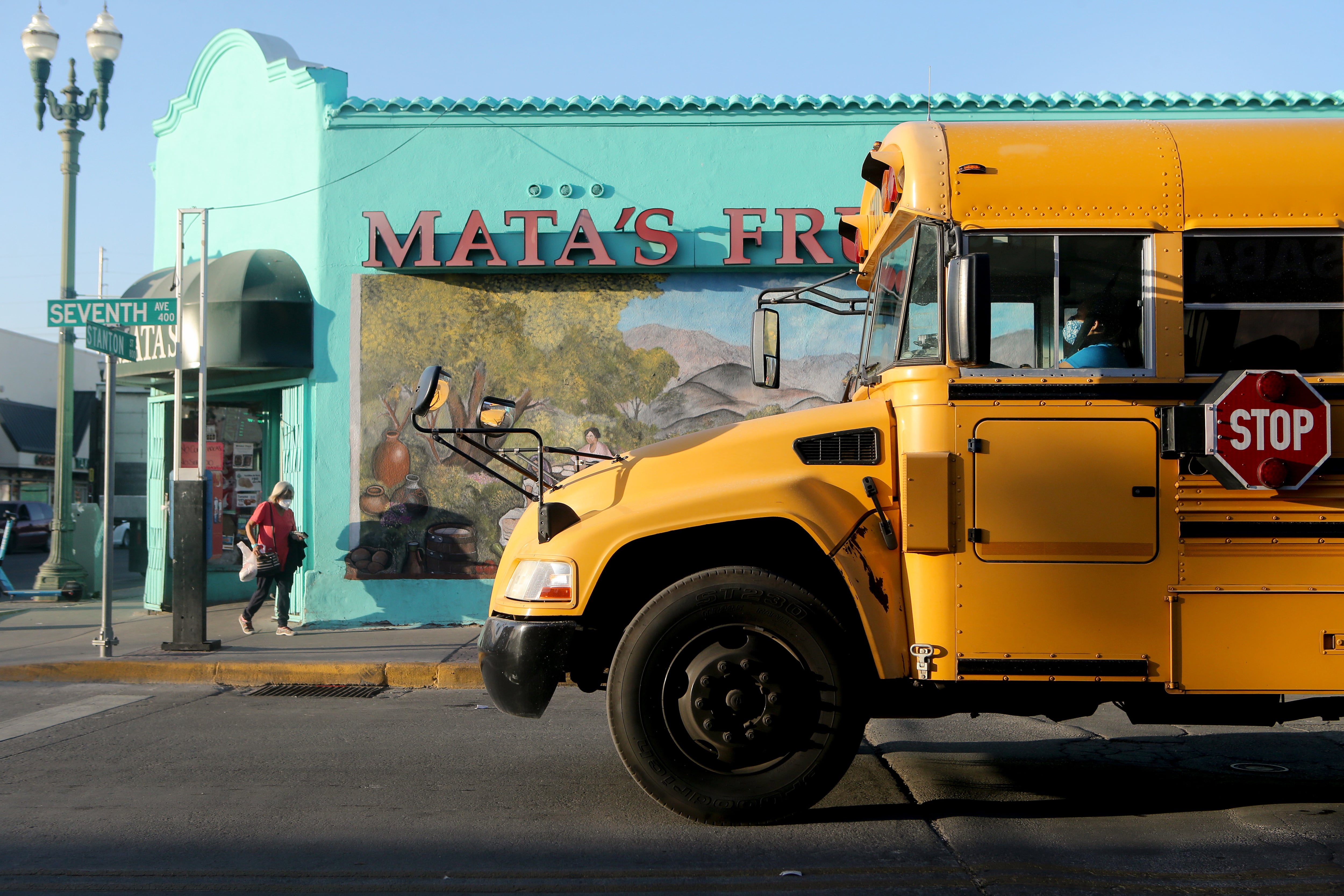 A yellow school bus with a masked driver drives up to an intersection in El Paso, Tex. There is a vibrant teal-colored storefront with a blonde woman wearing a pink shirt walking by on the corner of the street.