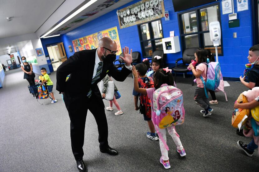Denver schools Superintendent Alex Marrero high-fives a young student wearing a pink backpack in a school hallway. The superintendent is wearing a suit and a face mask.