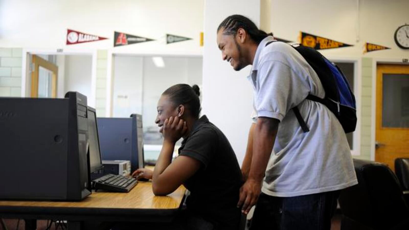 A woman sits at a desk looking at a desktop computer, while a man wearing a knapsack stands behind her also looking at the screen. Pennants of colleges and universities decorate a wall in the background.