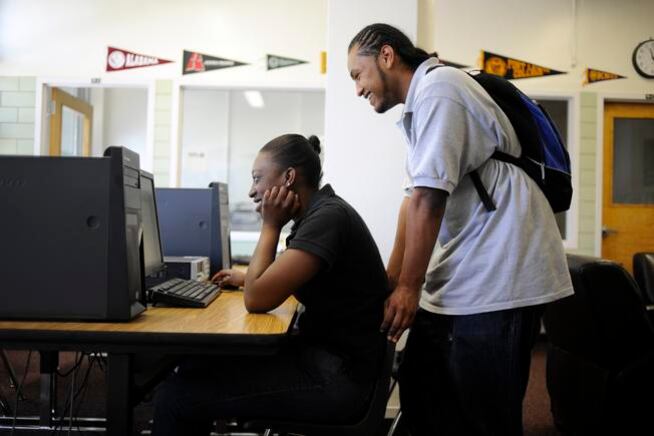 A woman sits at a desk looking at a desktop computer, while a man wearing a knapsack stands behind her also looking at the screen. Pennants of colleges and universities decorate a wall in the background.
