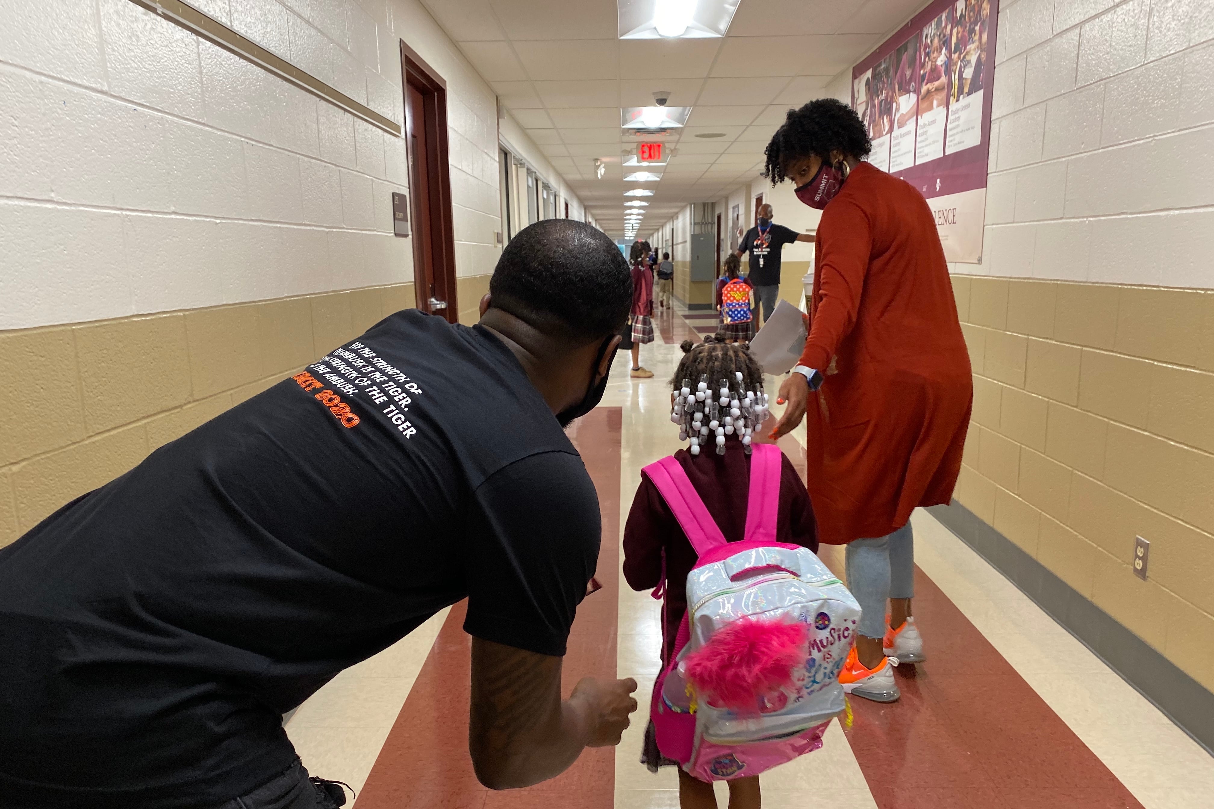 A masked woman in a long orange sweater guides a child carrying a pink backpack down a school hall, while a man in a black T-shirt bends down slightly behind the child.