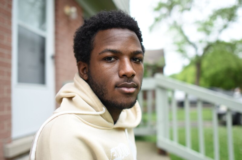 A young person with short dark hair and wearing a tan hoodie, poses for a portrait outside.