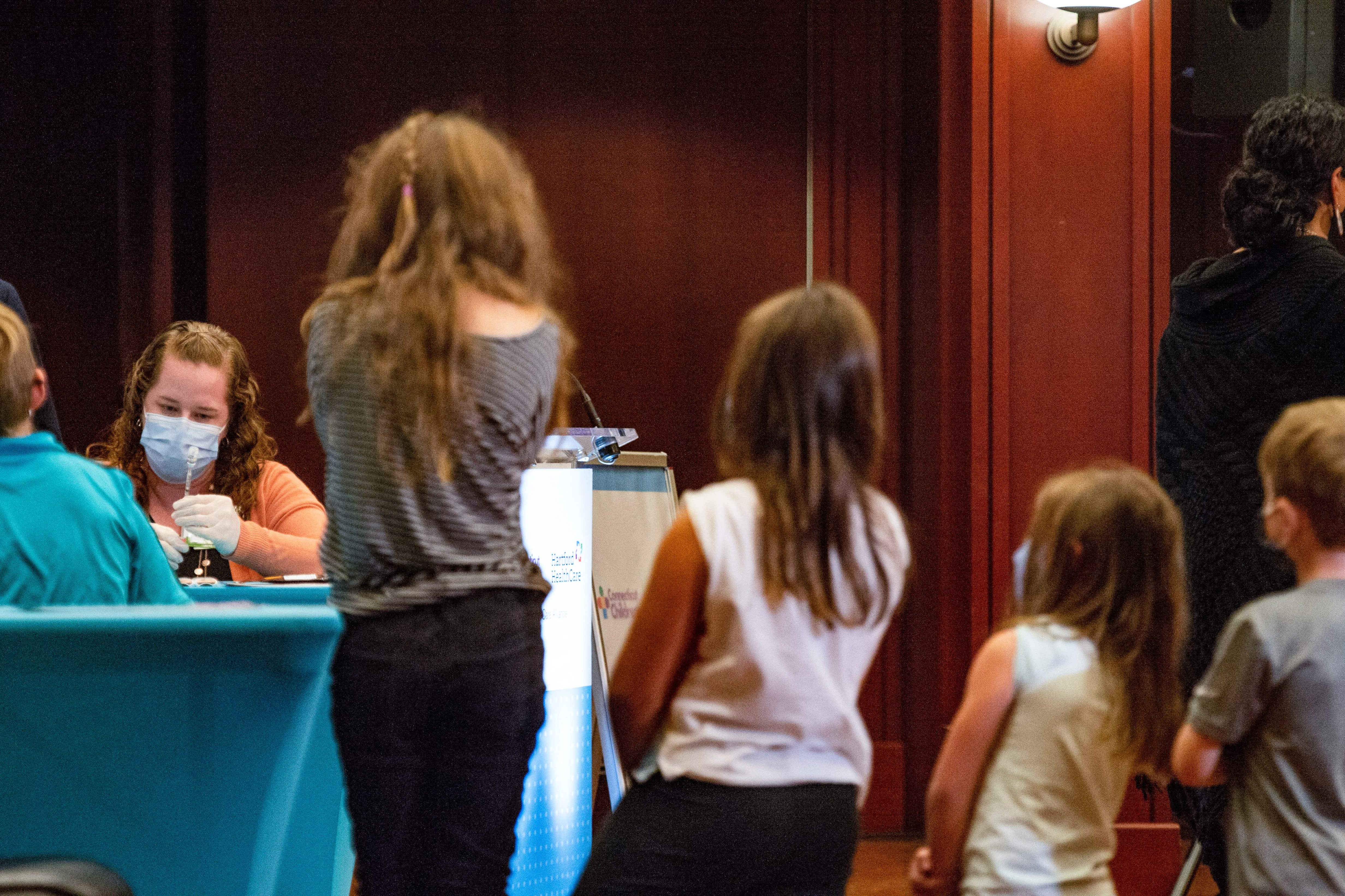 Young children wait in line as another child receives a dose of the COVID vaccine from a female healthcare professional.