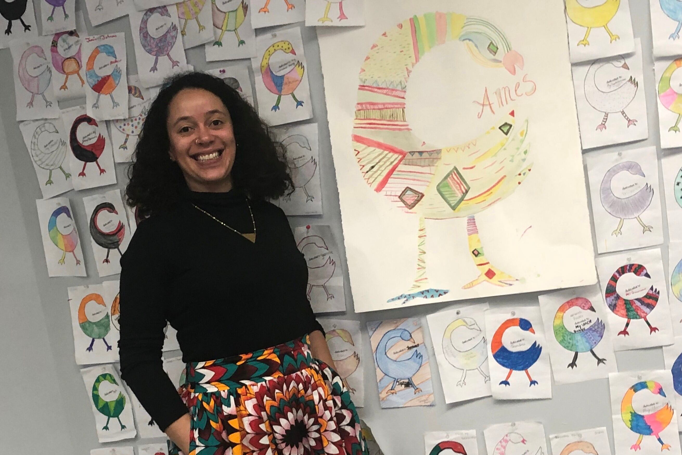 A teacher smiles, standing in front of a whiteboard wearing a black turtleneck.