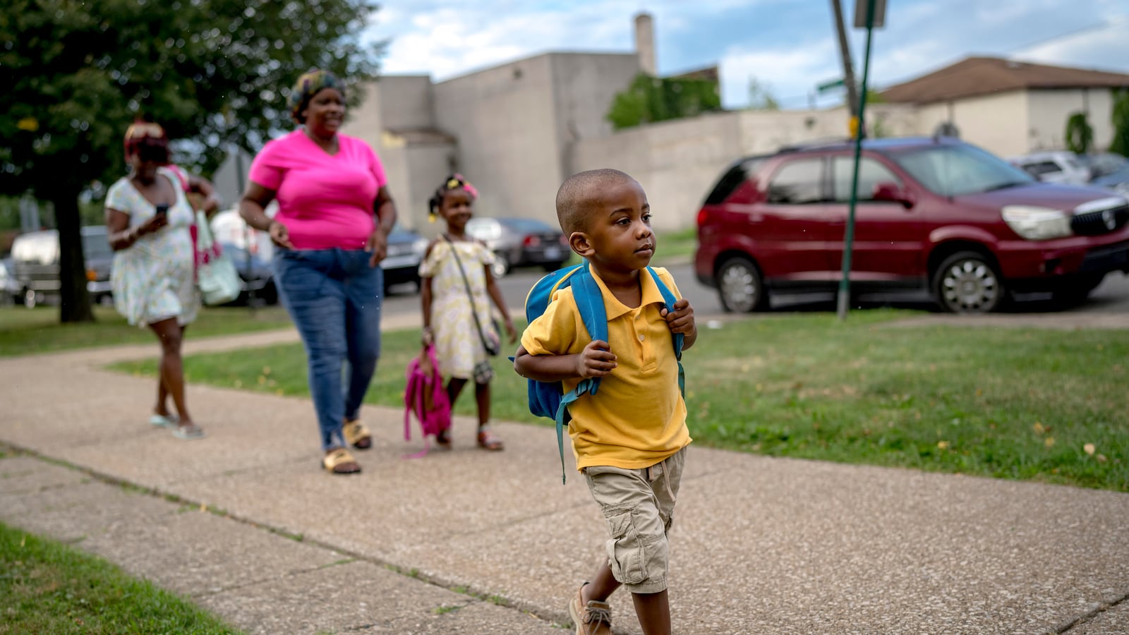 A child wearing a backpack walks toward a school building. Other children and parents can be seen behind him.