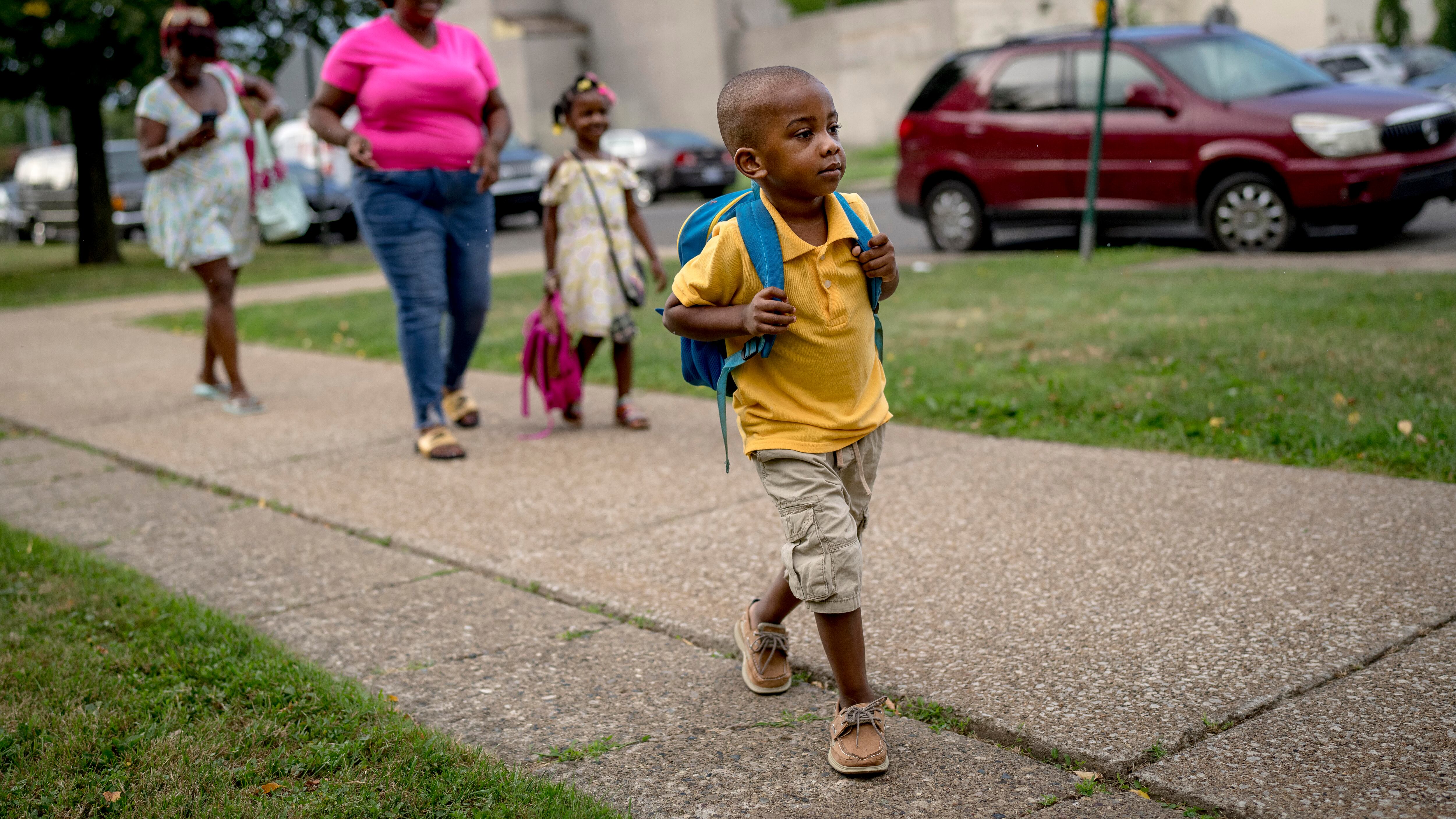 A child wearing a backpack walks toward a school building. Other children and parents can be seen behind him.