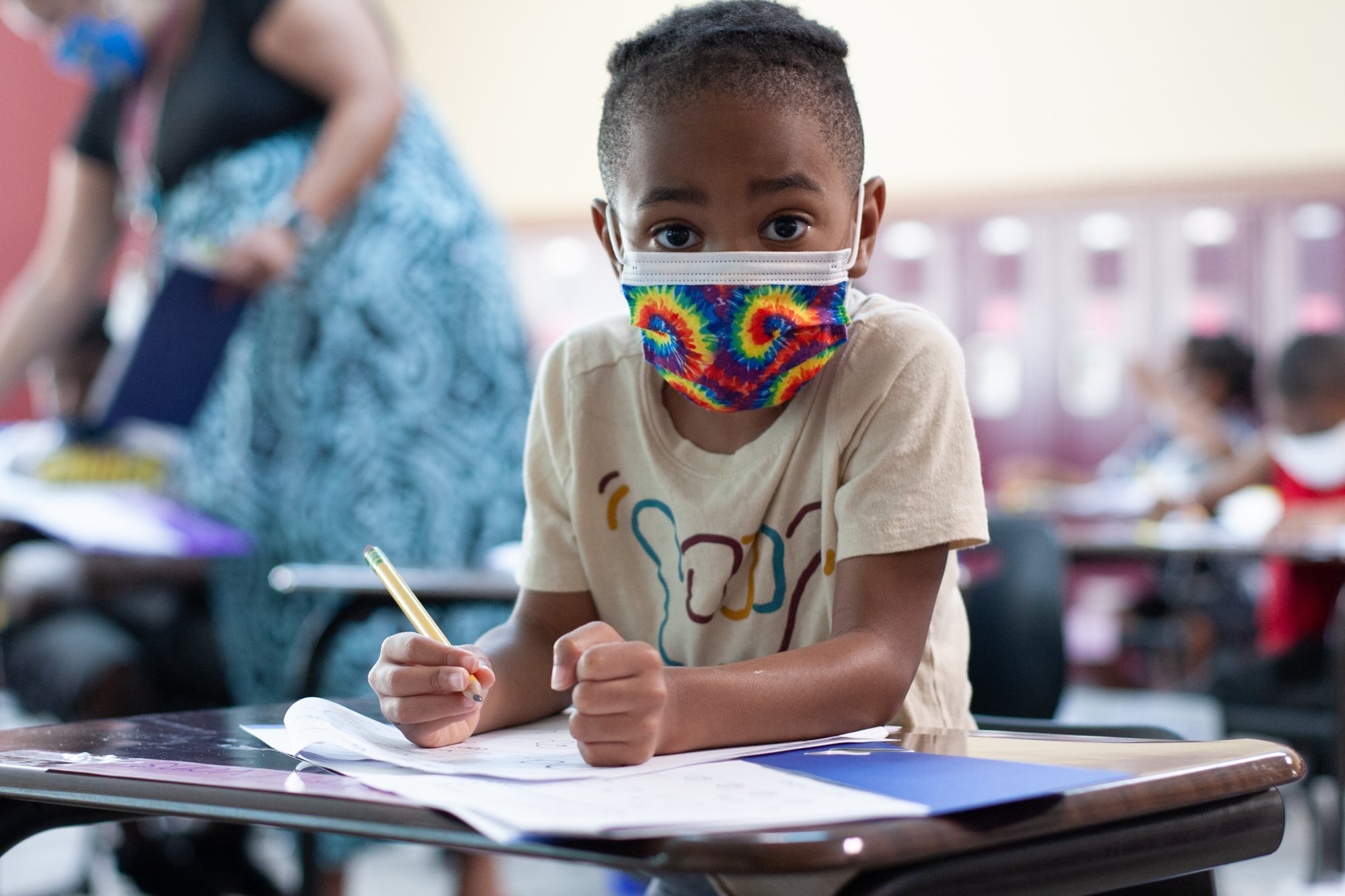A young boy in a beige shirt with a tie-dye mask looks up at the camera in the middle of writing on a worksheet with a pencil as a teacher helps a student in the background
