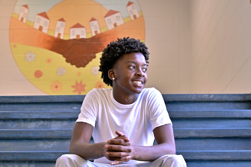 A high school student with dark hair and wearing a white t-shirt sits and smiles for a portrait on a set of stairs in a school.