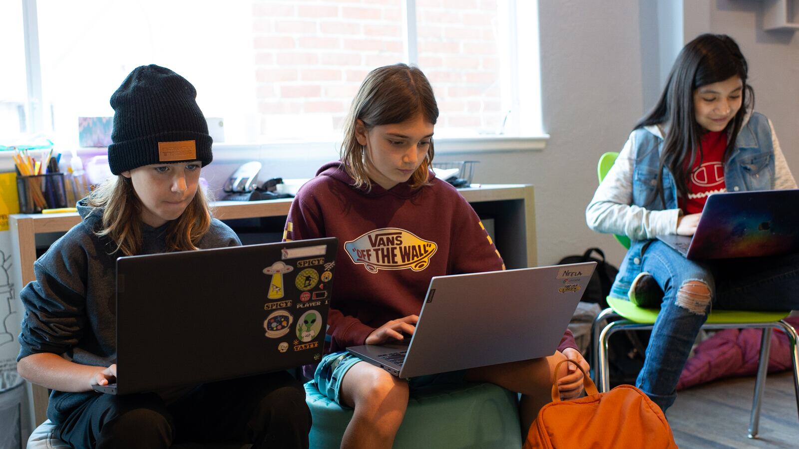 Three children work intently on laptops. Two are sitting on what look like ottomans or cushions, and the third is in a chair.