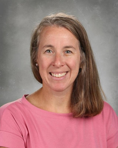 A photograph of a white woman with long blonde hair and wearing a pink shirt in front of a grey backdrop.