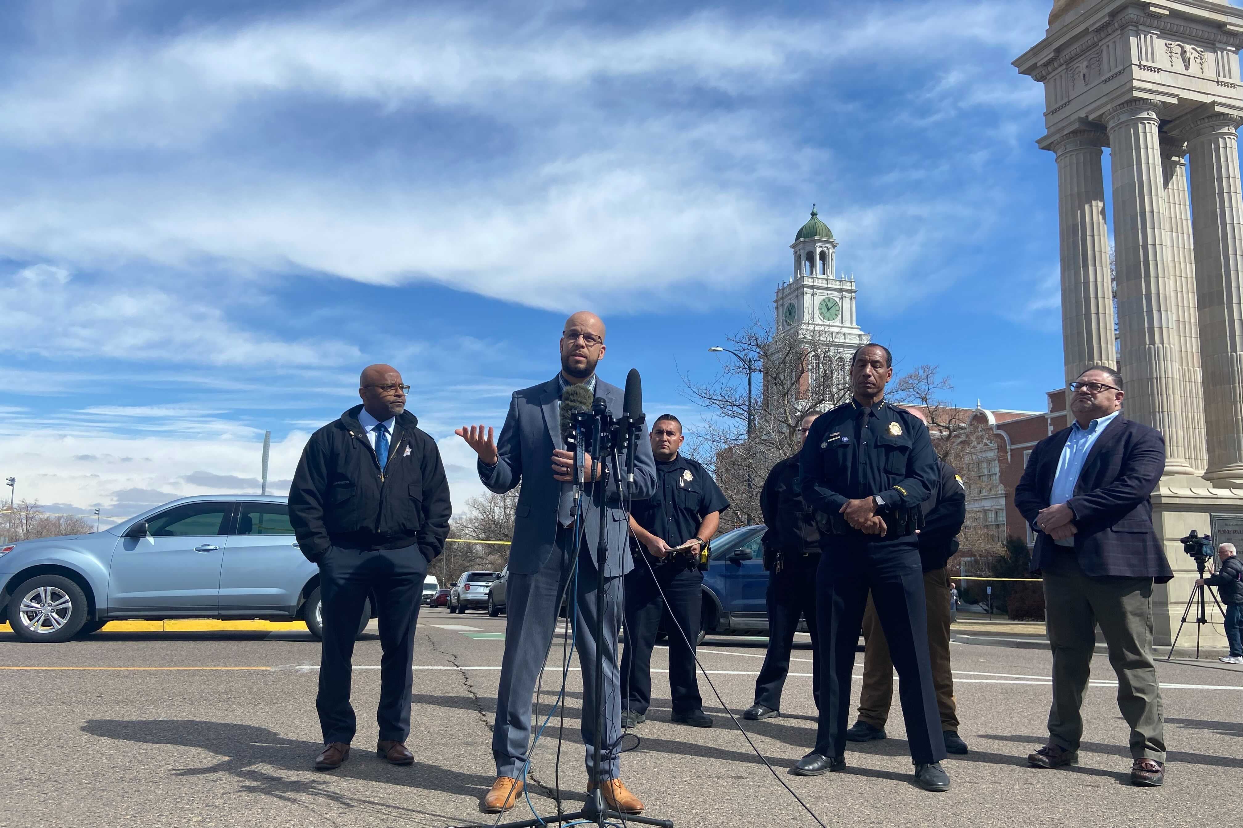 A group of men in suits and one in police dress uniform speak at a press conference. They are outside with a podium and microphones set up in front of them. East High School is visible behind them.