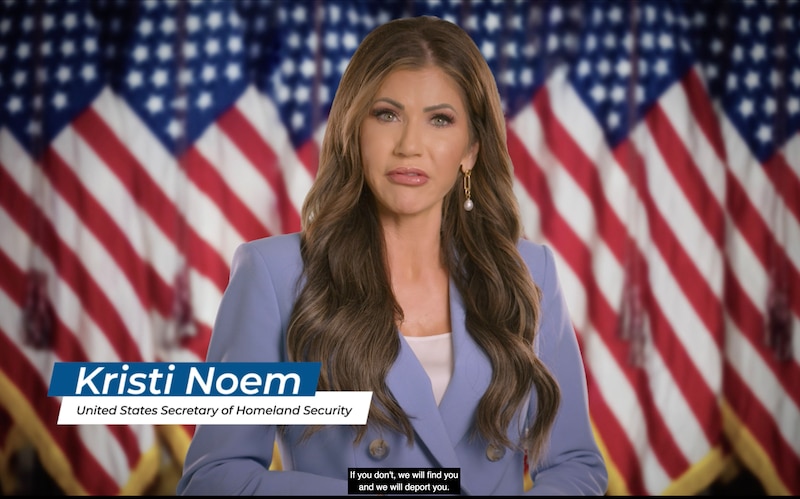 A screenshot of a white woman with long brown hair wearing a light purple suit standing in front of a row of American flags.