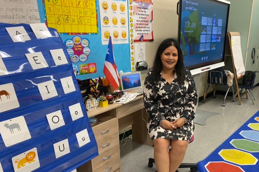 A teacher smiles in a classroom where students’ artwork dots the walls and a colorful play mat is on the floor.