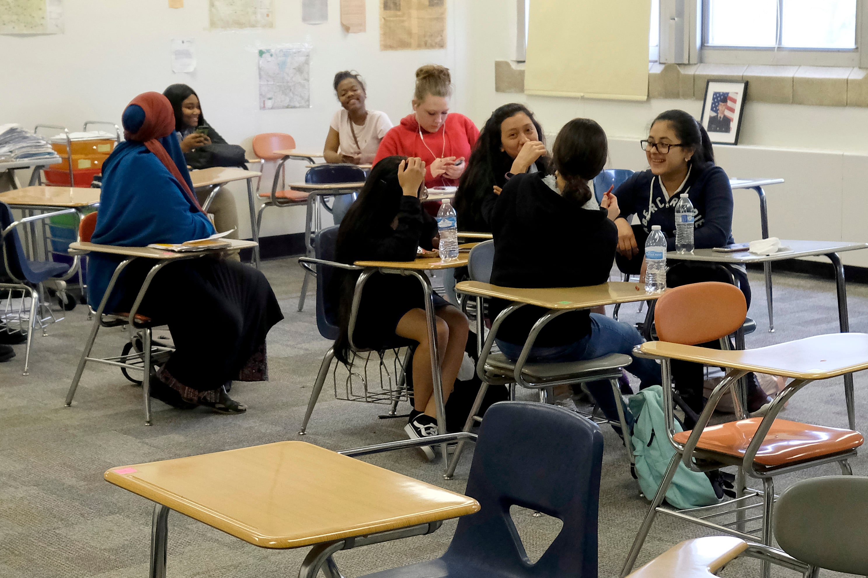 Eight high school students sit at desks in a classroom with a window in the background.