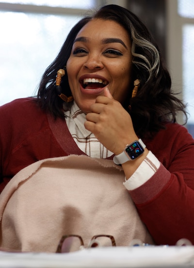 A woman with long dark hair and wearing a red sweater sits at a desk in a classroom.