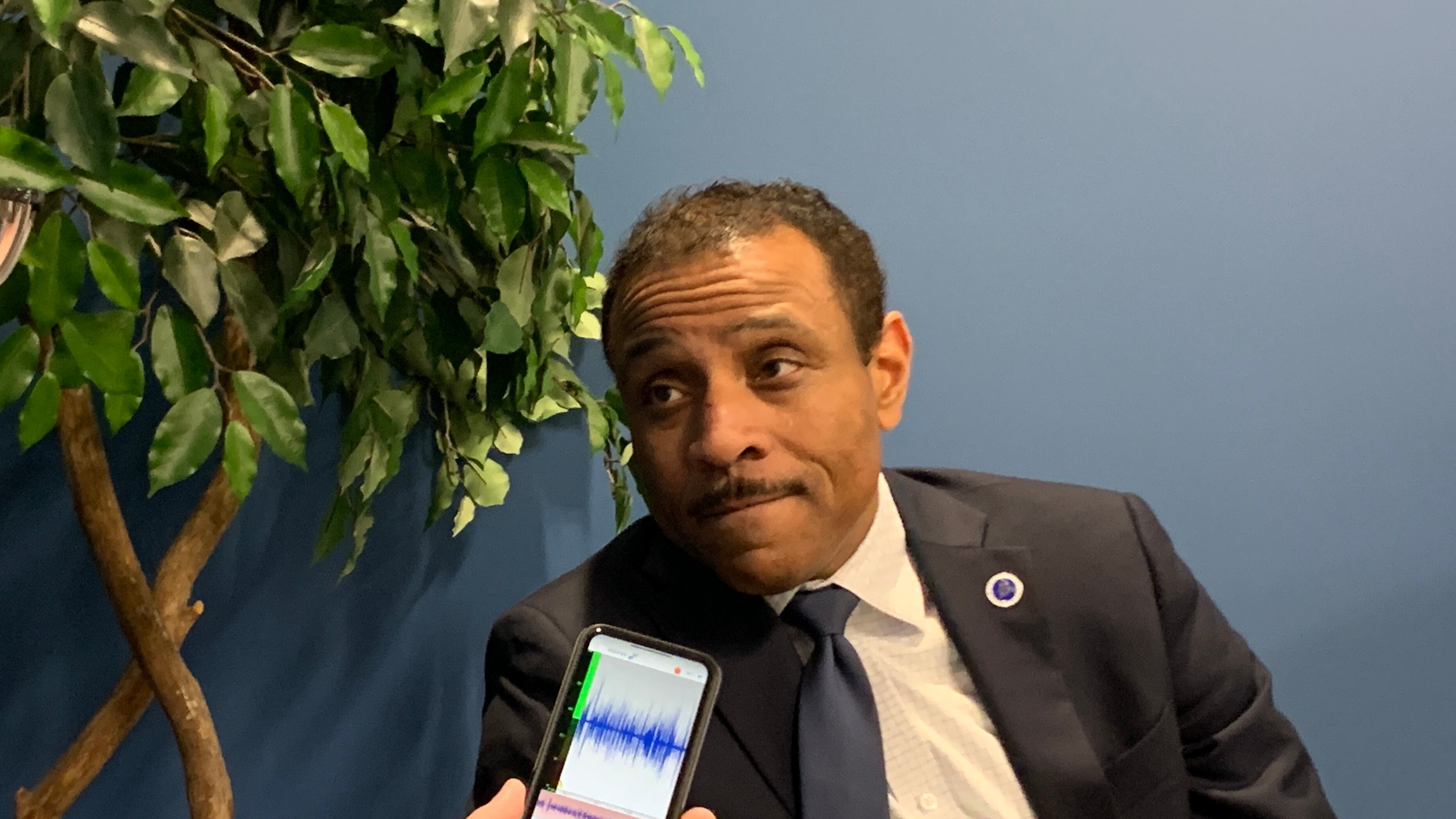 A man in a dark suit and white shirt leans to his right while a person holds a phone near his face in front of a plant a blue wall.